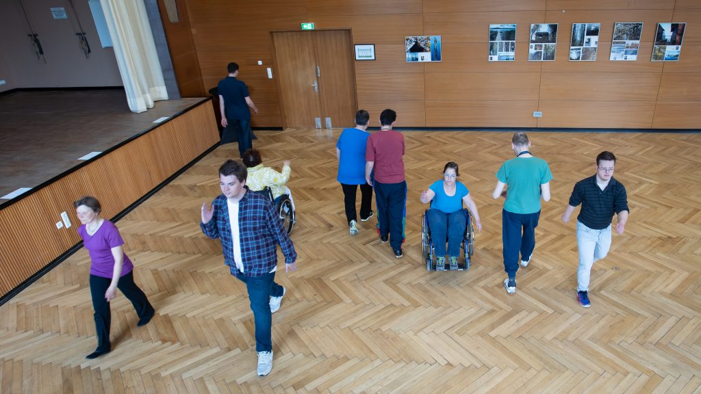 Draufsicht auf acht Tänzerinnen und Tänzer, die durch den Raum schreiten und rollen