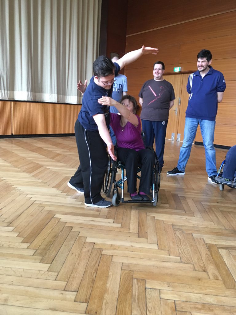 Eine Tänzerin im Rollstuhl deutet eine Hebefigur mit einem anderen Tänzer an